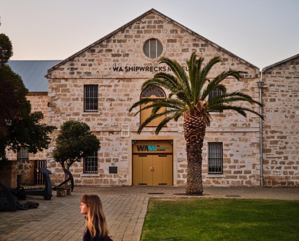 A limestone brick building with a large wooden door and windows behind a palm tree.