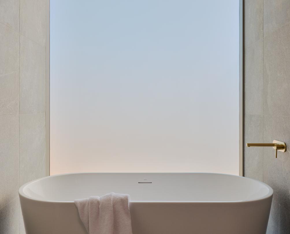 A deep bath in front of a floor to ceiling frosted window.