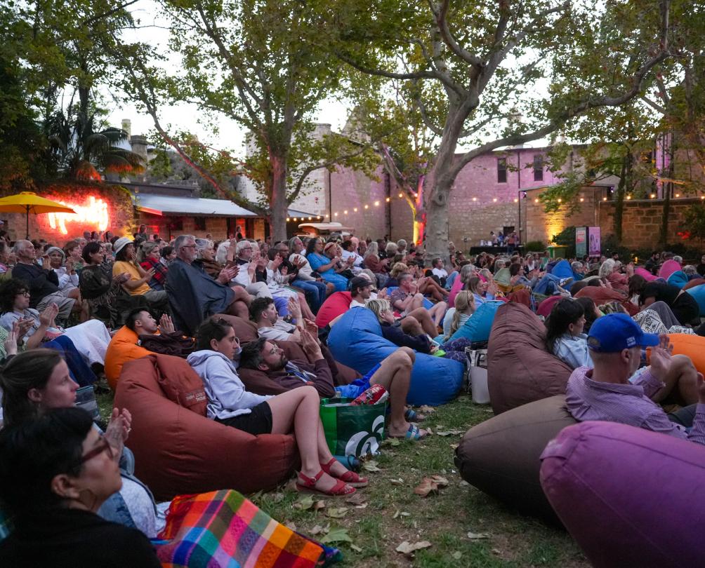 A large group of people sit on bean bags on a lawn watching a film on a big screen.
