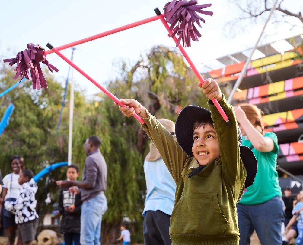A smiling young child holds two pink batons holding up a third pink baton with tassels on the end.