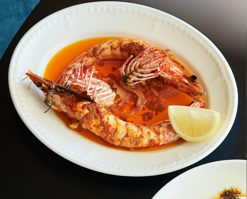 A dish of grilled prawns with a wedge of lemon on a table.