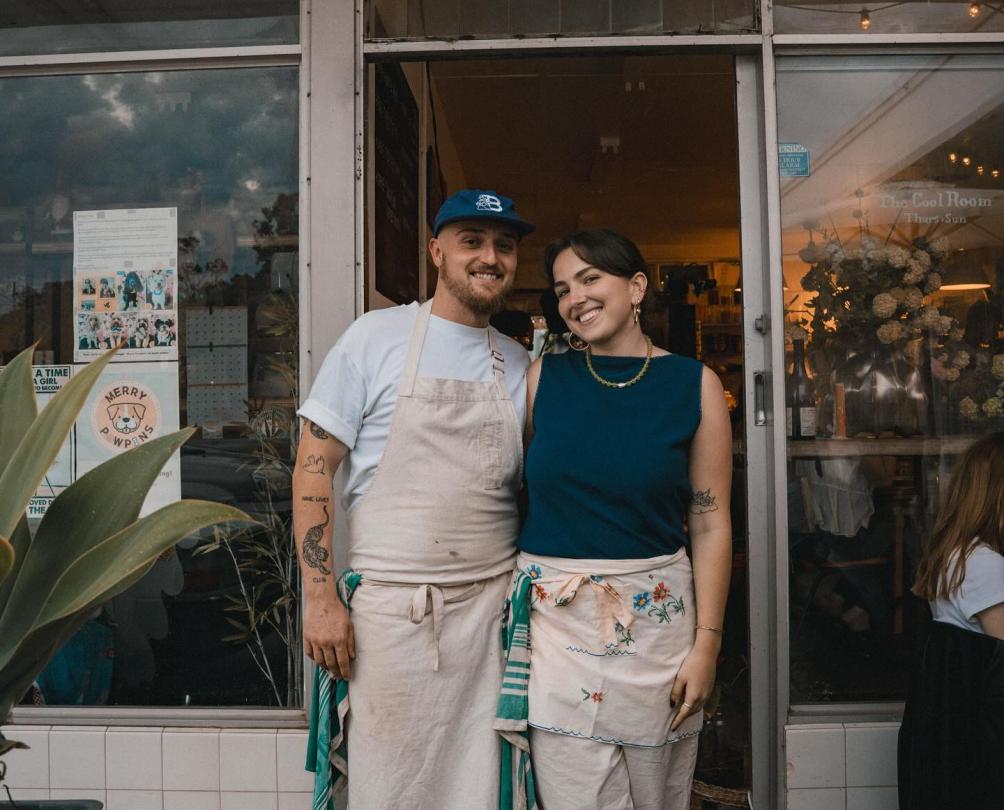 A man and a woman wearing aprons standing outside the front door of a cafe smiling for the camera.