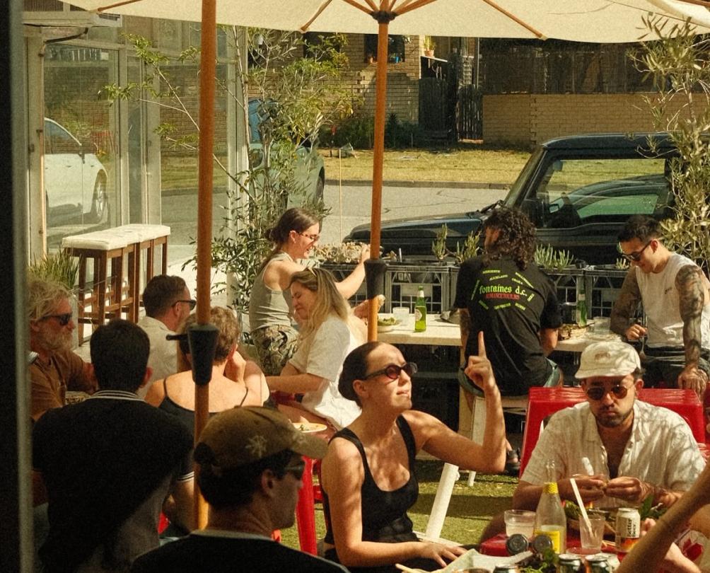 The exterior of a cafe with big white umbrellas and people dining under them on a sunny day.
