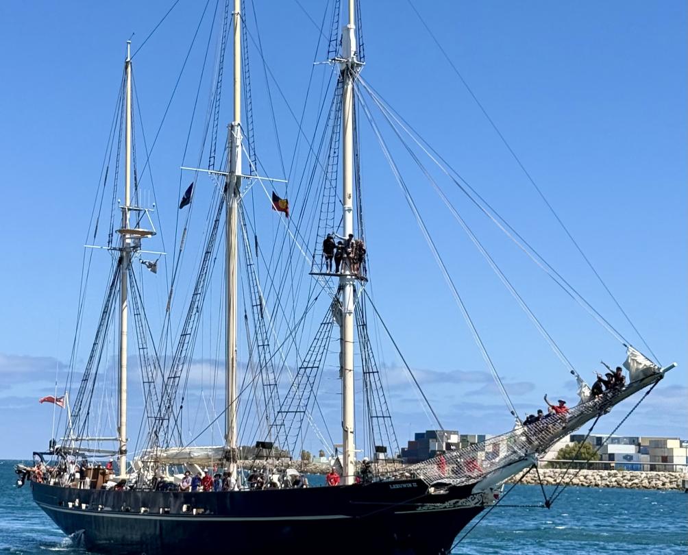 A tall ship with three masts and its sails down heading into a harbour with a large group aboard.