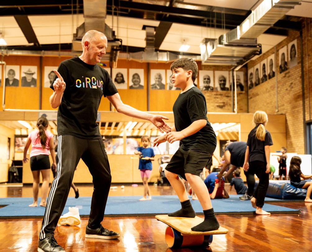 A young boy and a man practice circus tricks in a hall.