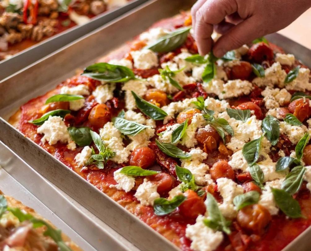 A hand placing fresh basil on a square pizza in a tray.