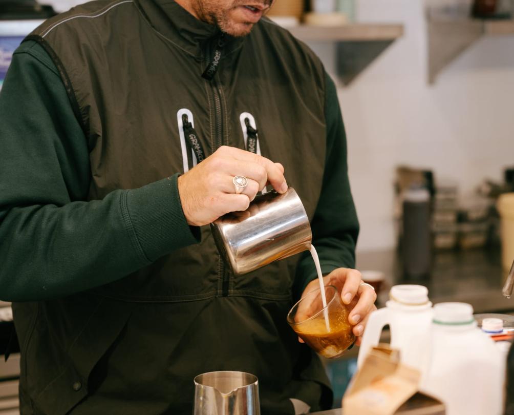 a man pouring a coffee in a cafe with milk coming out of the jug into a cup