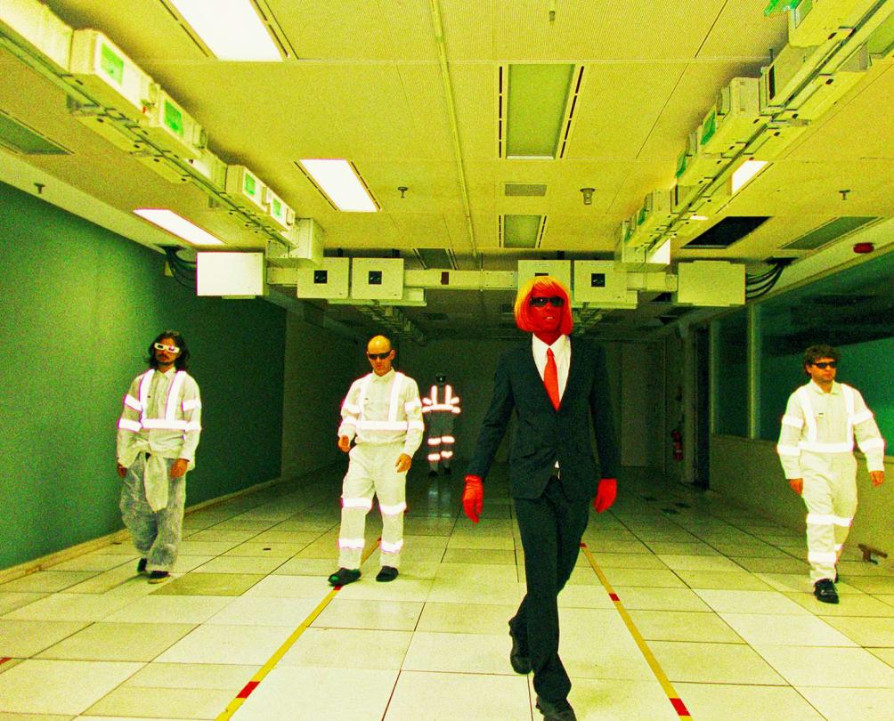 a man with red skin and 3 other men in white jumpsuits in a weird room 