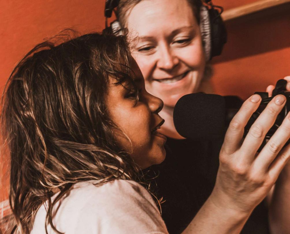 a girl talking into a microphone with another woman looking on in the background