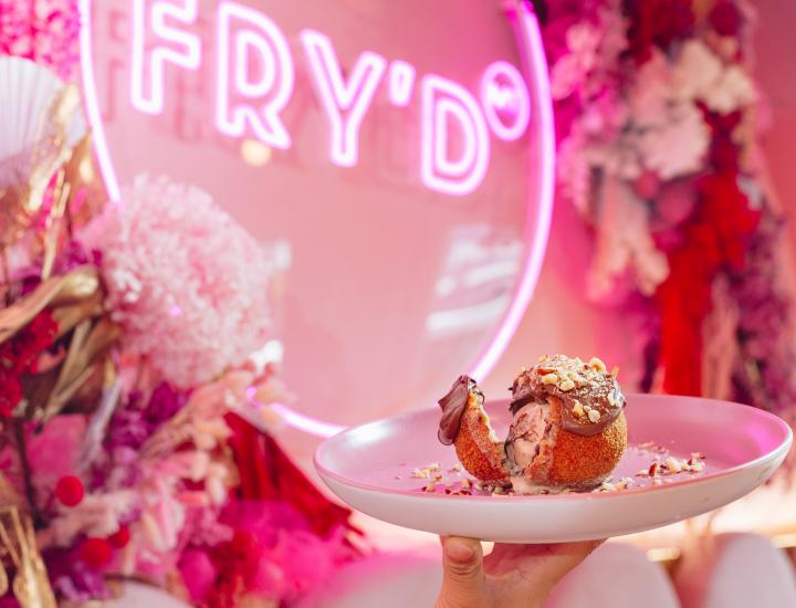 hand holds white plate with a bowl of fried ice cream covered in chocolate in front of a bright pink background and pink neon sign saying FRY'D TM
