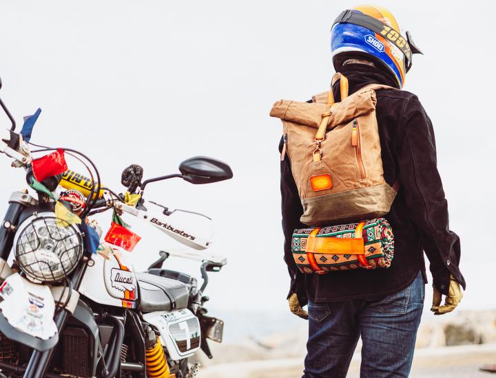 person wearing black jacket, motorcycle helmet and brown backpack stands next to motorbike