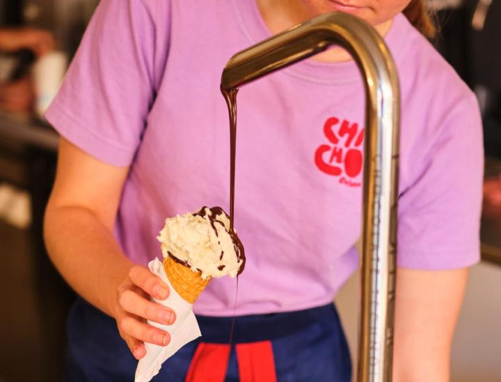 person in pink ChiCho shirt drizzles chocolate sauce over an ice cream in a a waffle cone 