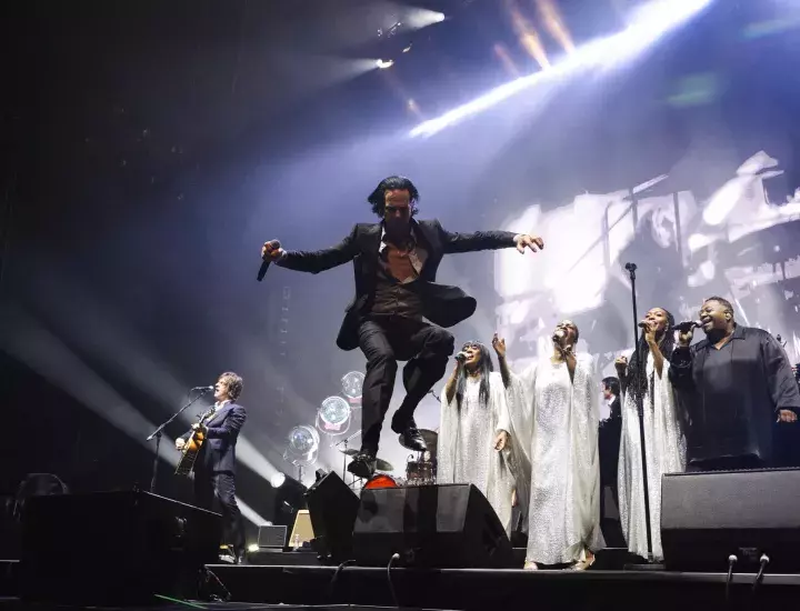 A musician dressed in a black suit jumps off a stage with a choir behind him.