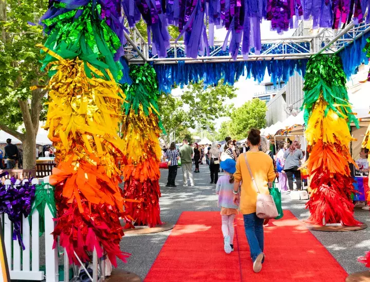 Two people walk underneath a rainbow coloured marquee on a red carpet.