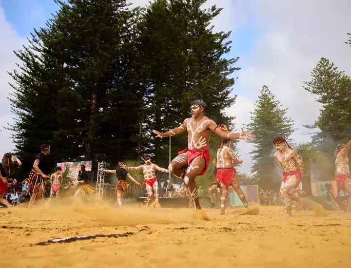 A group of young indigenous Australians perform a traditional cultural dance on sand.