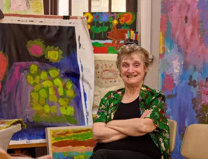 A woman with arms folded, smiling in front of a group of bright paintings.