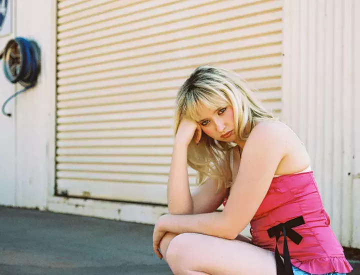 A blonde woman in a pink dress crouching down in front of a cream roller door.