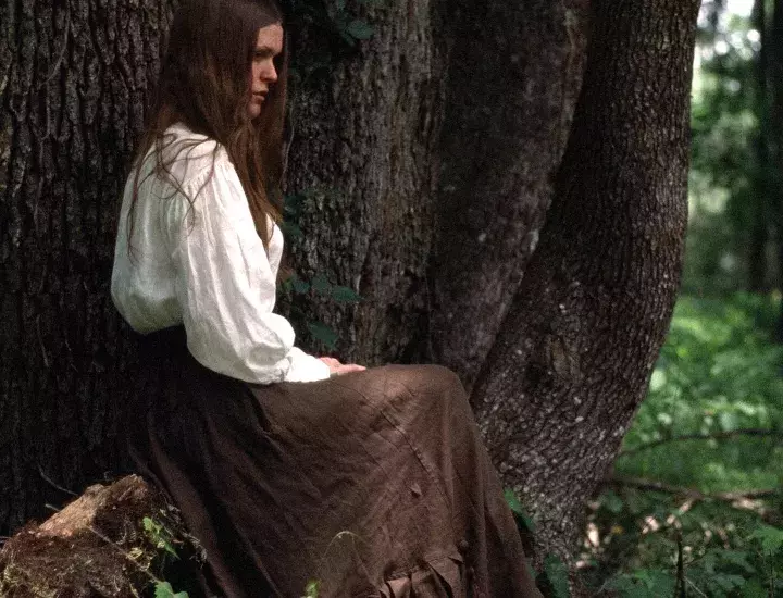 A woman in a long white shirt and brown skirt sits under a tree in a forest.