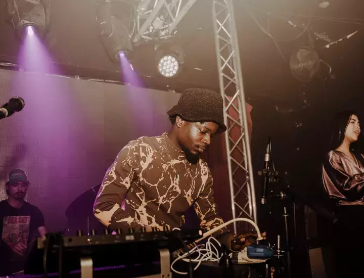 A man stands behind a DJ booth wearing a printed shirt and a felt hat.