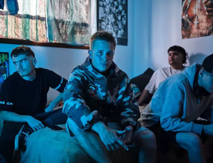Four young men sitting on a bed in a bedroom in blue light.