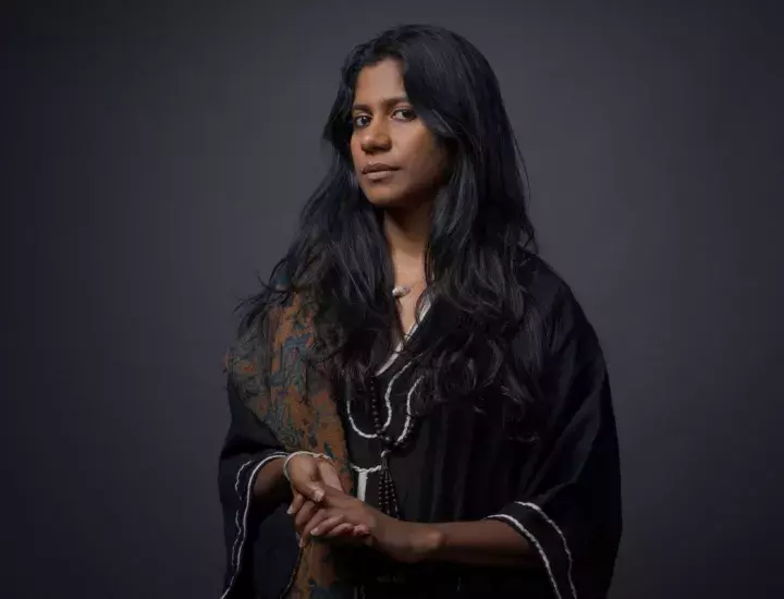 A woman with long dark hair wearing an Indian shawl and jewellery stares intently into a camera.