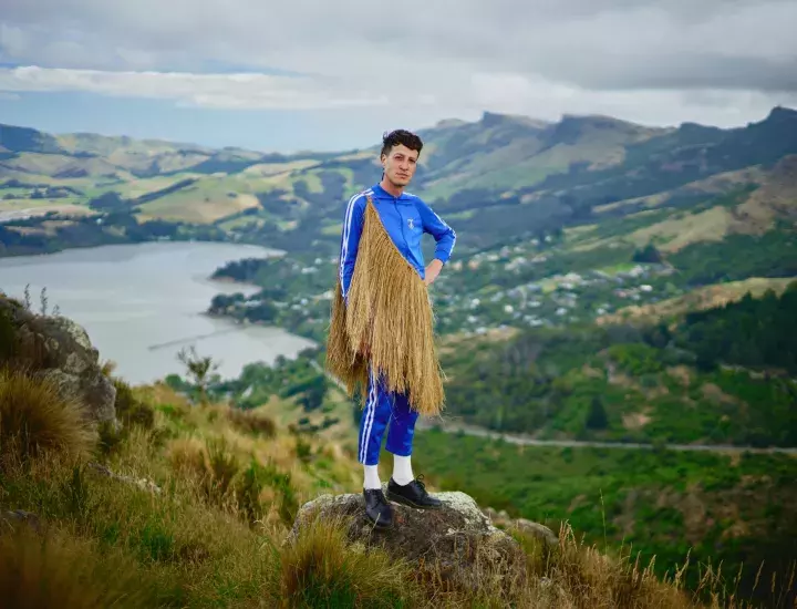 A man in a blue tracksuit, black dress shoes, and traditional Maori woven cape stands on top of a mountain with a valley behind him.