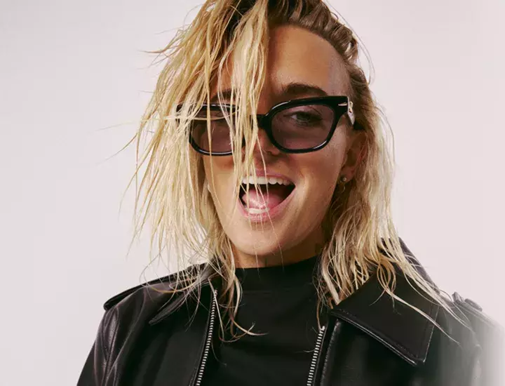 A woman with messy short blonde hair wearing a leather jacket and pulling a face at the camera.