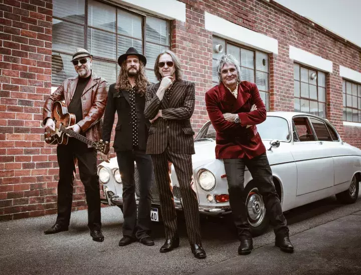 Four men  in 70s era clothing stand in front of a white 70s era car.