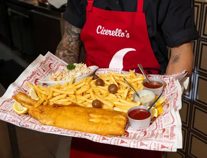a big plate of fish and chips with different sauces being held by a man with a red apron